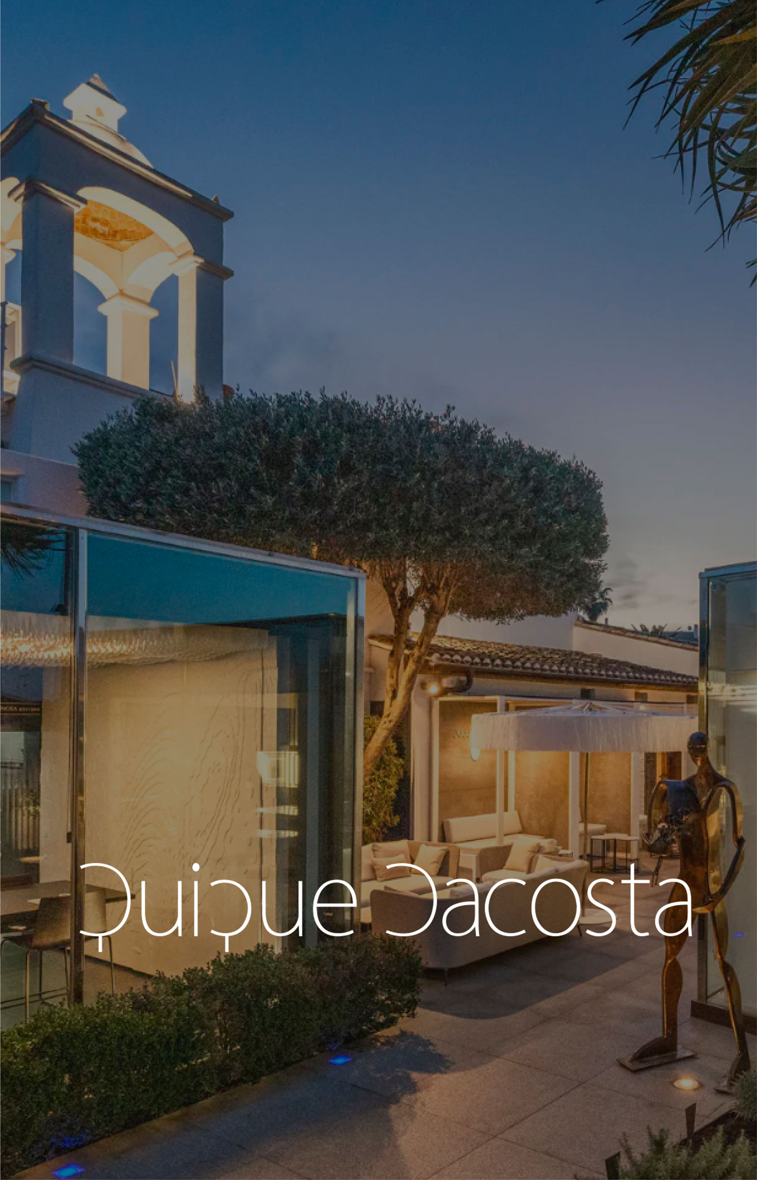 Exterior de Quique Dacosta al anochecer, con arquitectura blanca, terraza, vegetación y una escultura.